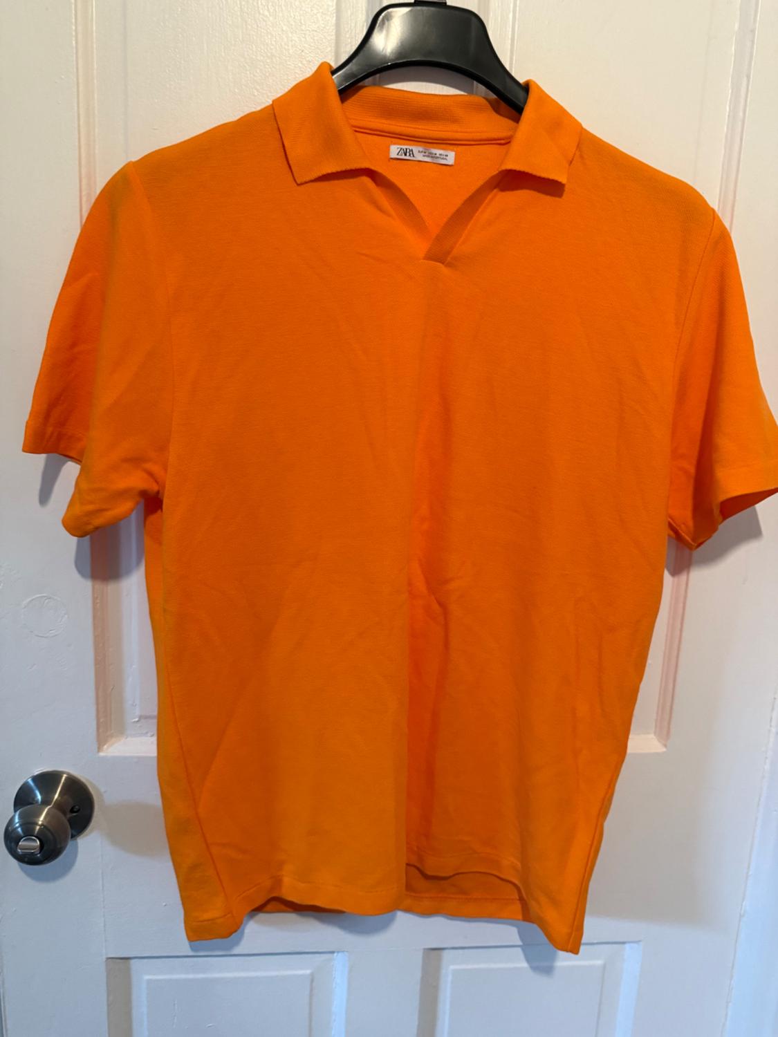 Orange polo shirt hanging on a black hanger over a white door. Visible tag on the collar.