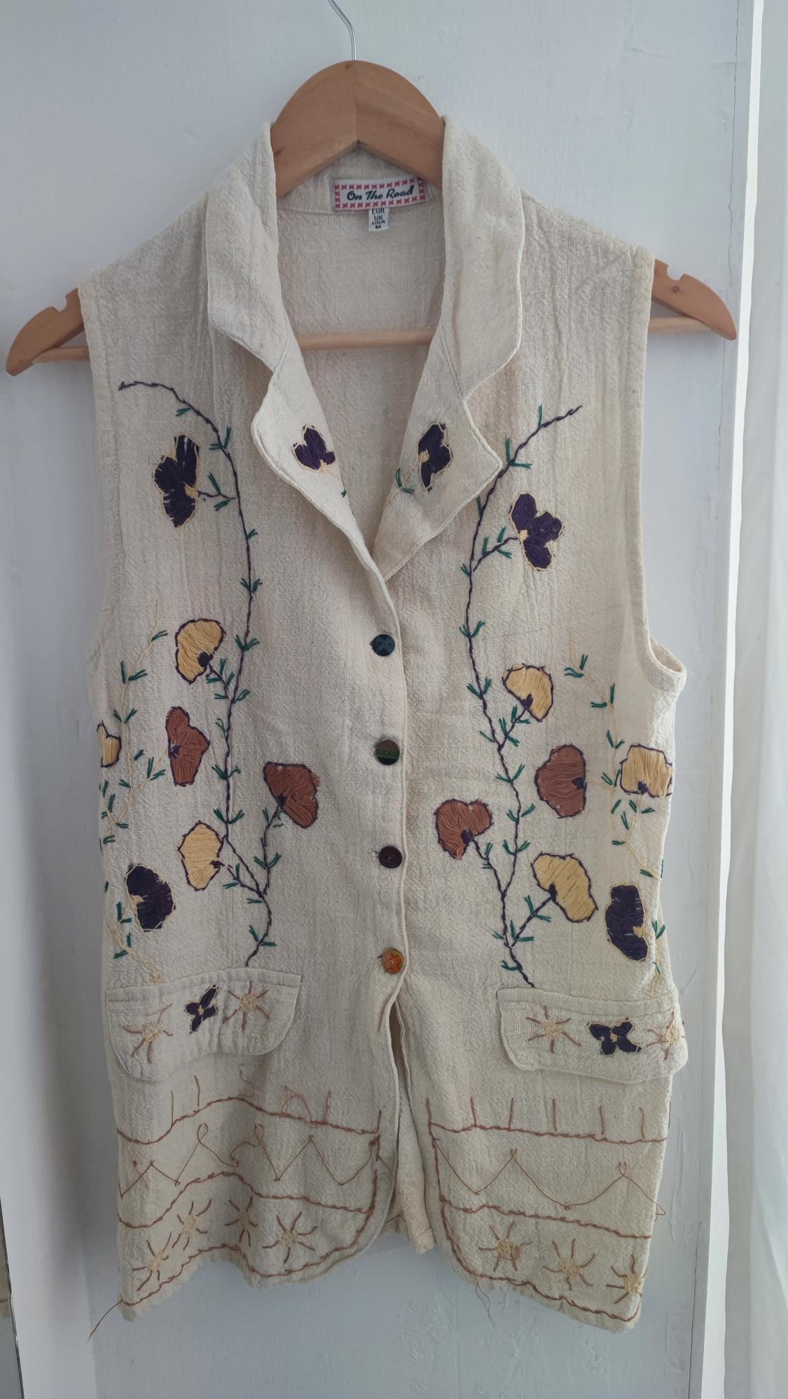 Beige waistcoat hanging on a hanger, with floral embroidery in shades of purple, yellow, and brown. "On The Road" label visible.