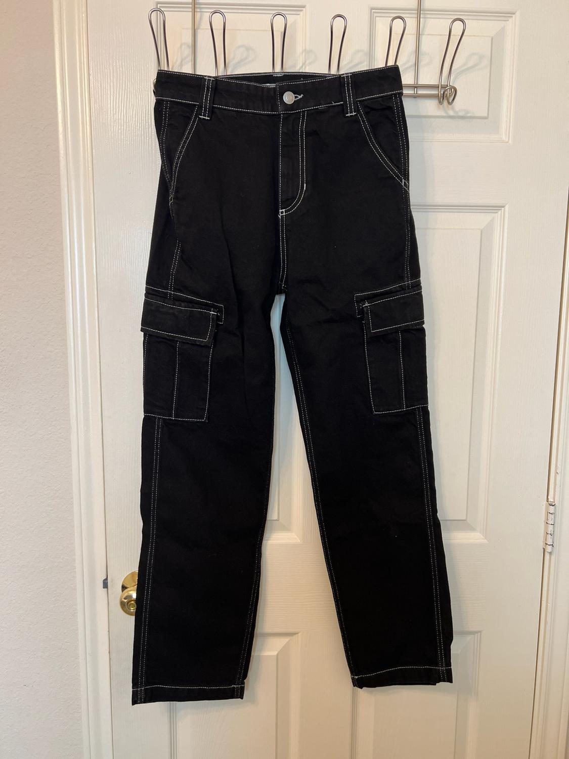Black cargo trousers with contrasting white stitching, hanging on a white interior door.