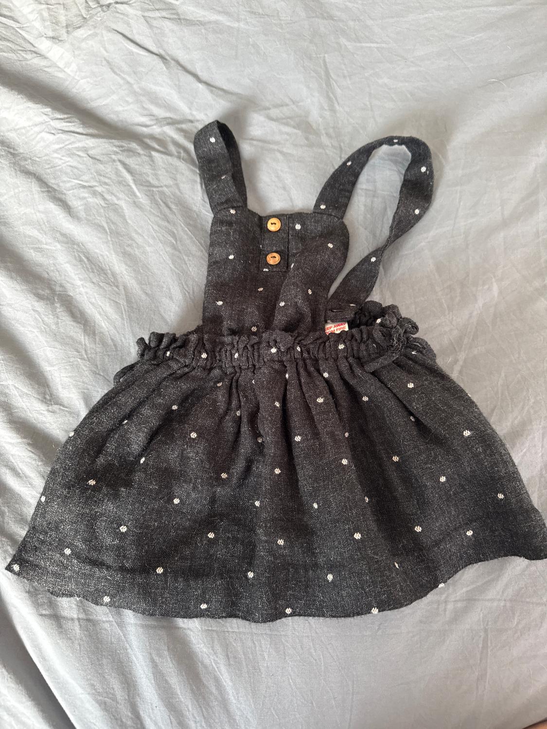 Dark grey children's dress with straps, wooden buttons, and a white polka dot print on a light grey crinkled fabric.