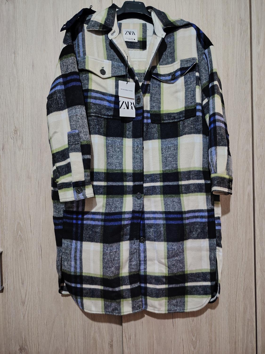 Checked coat in black, blue, and green tones, hanging on a hanger. Visible label with the text "ZARA". Light wooden background.