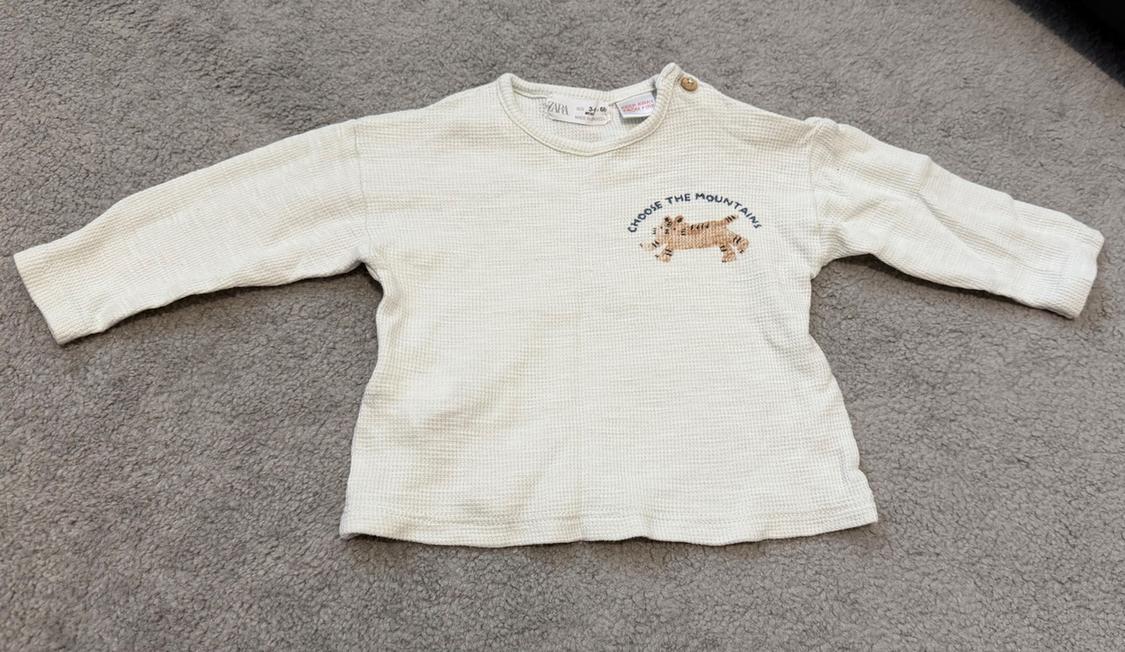 Beige long-sleeved baby jumper on a grey rug, with the text "Choose the mountain" and a leopard illustration on the chest.