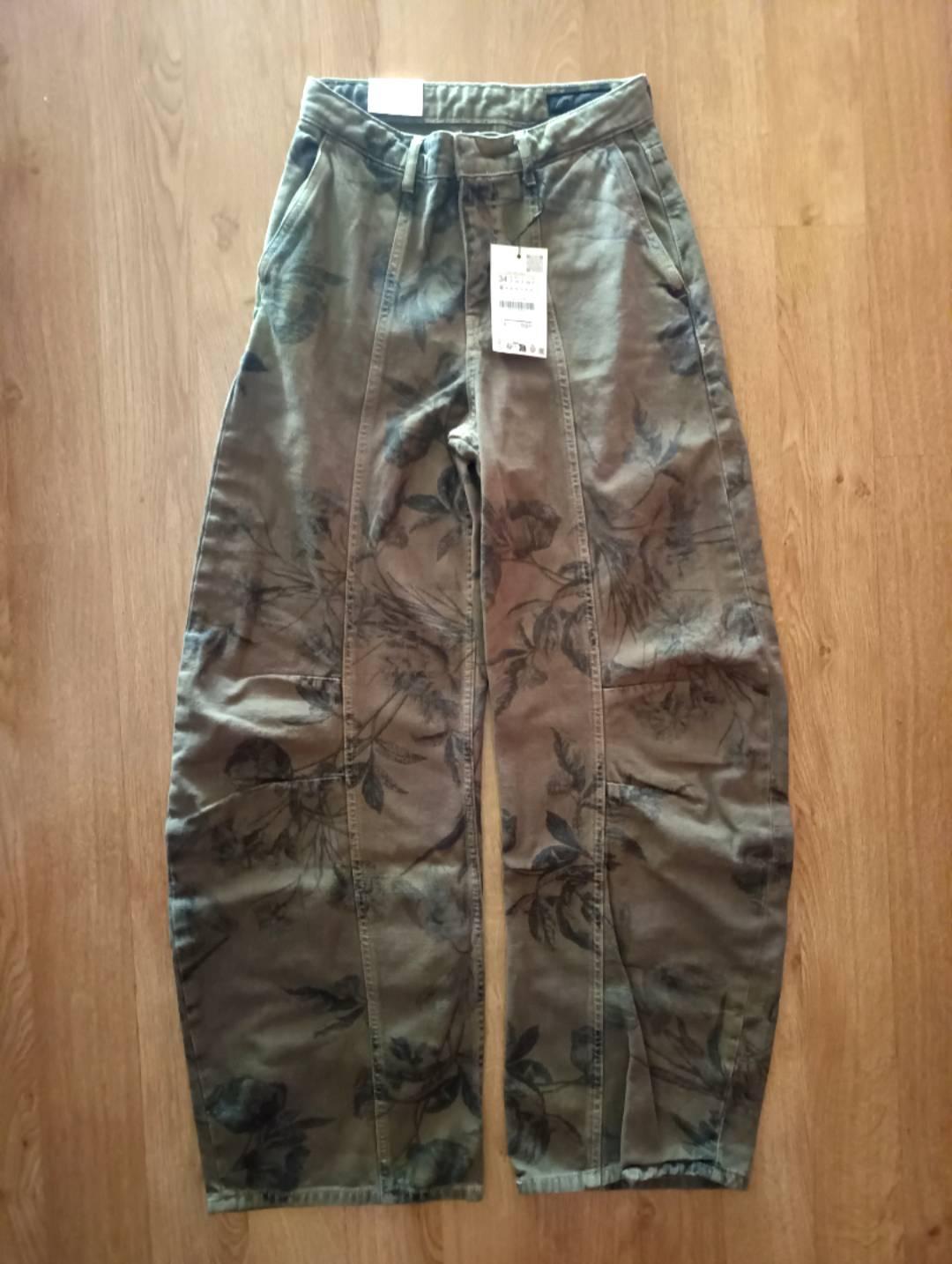 Wide green trousers with a dark floral print, laid out on a wooden floor. There is a label on the waistband.
