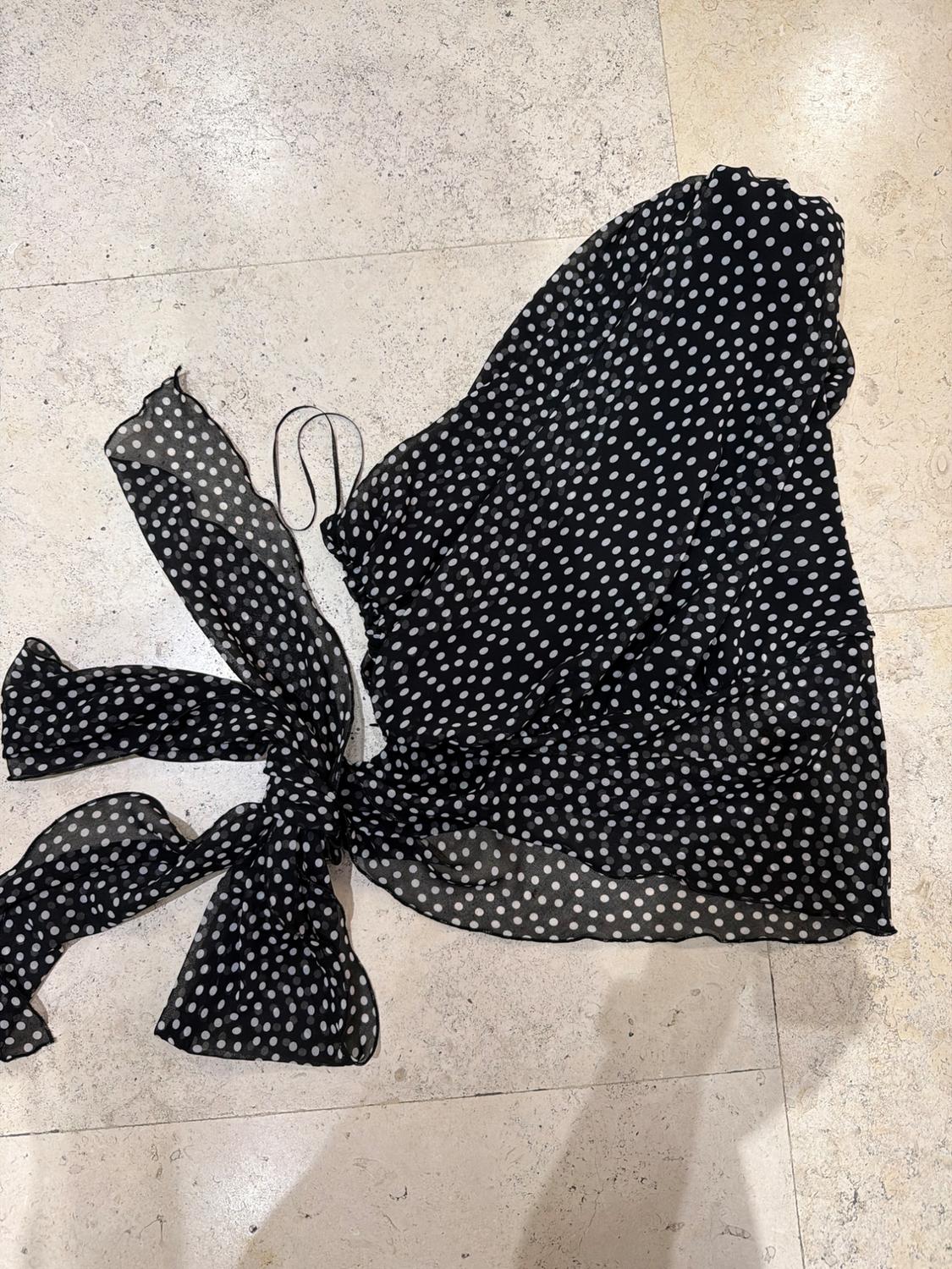 Black polka-dot camisole with thin straps and a large bow detail, made from a fluid fabric, on a light stone floor.