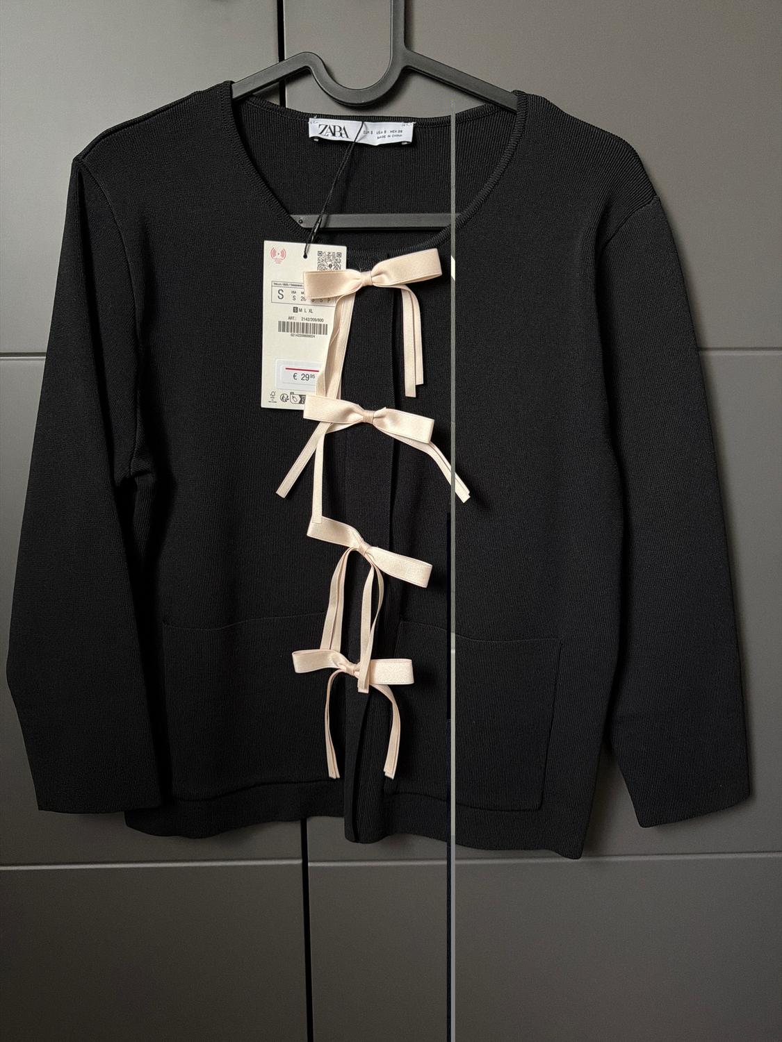 Black jacket with a beige bow on the front, hanging on a hanger. Visible tag with a price of €29.95. Grey background.