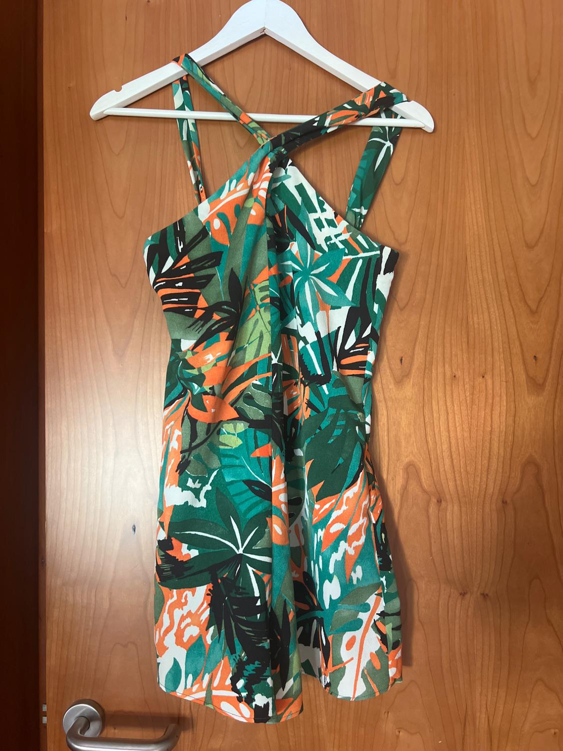 Short dress with a tropical print in shades of green, orange and white, with crossover straps, hanging on a hanger over a wooden door.