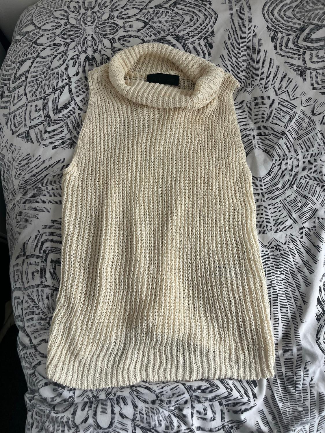 Sleeveless high-neck knitted top in cream, against a fabric background with a black-and-white print.