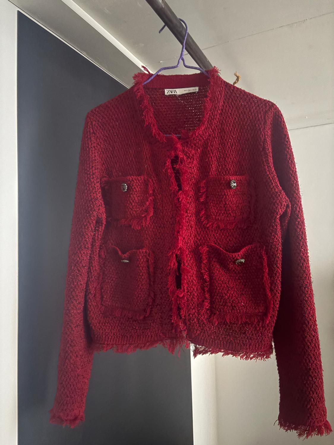 Red knitted jacket with pockets and buttons, hanging on a purple hanger. Visible label with text "ZARA". Dark background.