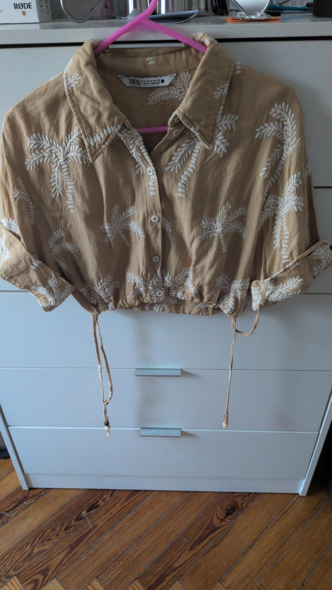 Mustard-coloured cropped shirt with white palm trees and ties, hanging on a pink hanger in front of a white chest of drawers. Text: RØDE, ZARA.
