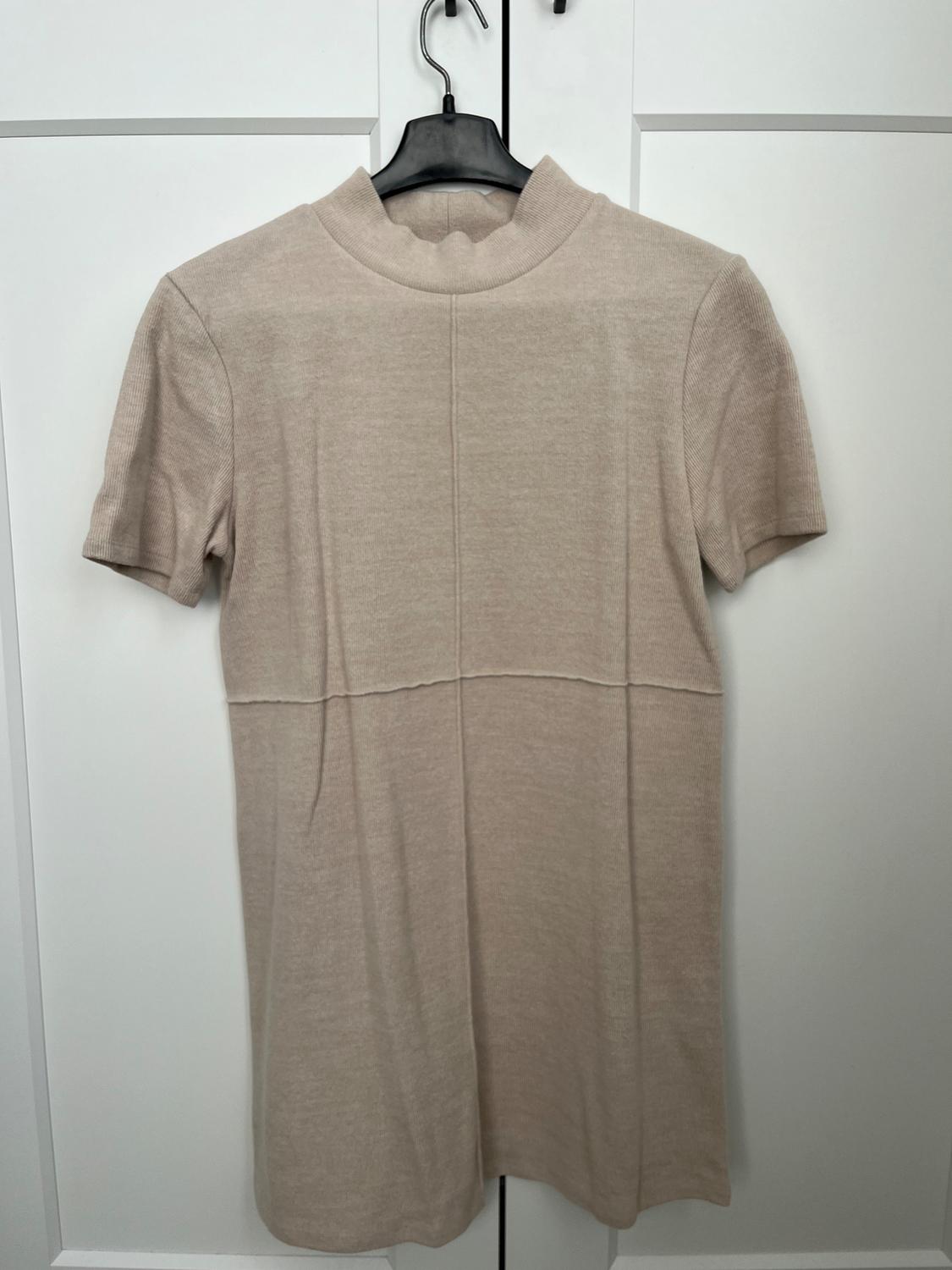 Beige short-sleeved, high-neck dress on a black hanger, hanging in front of a white door.