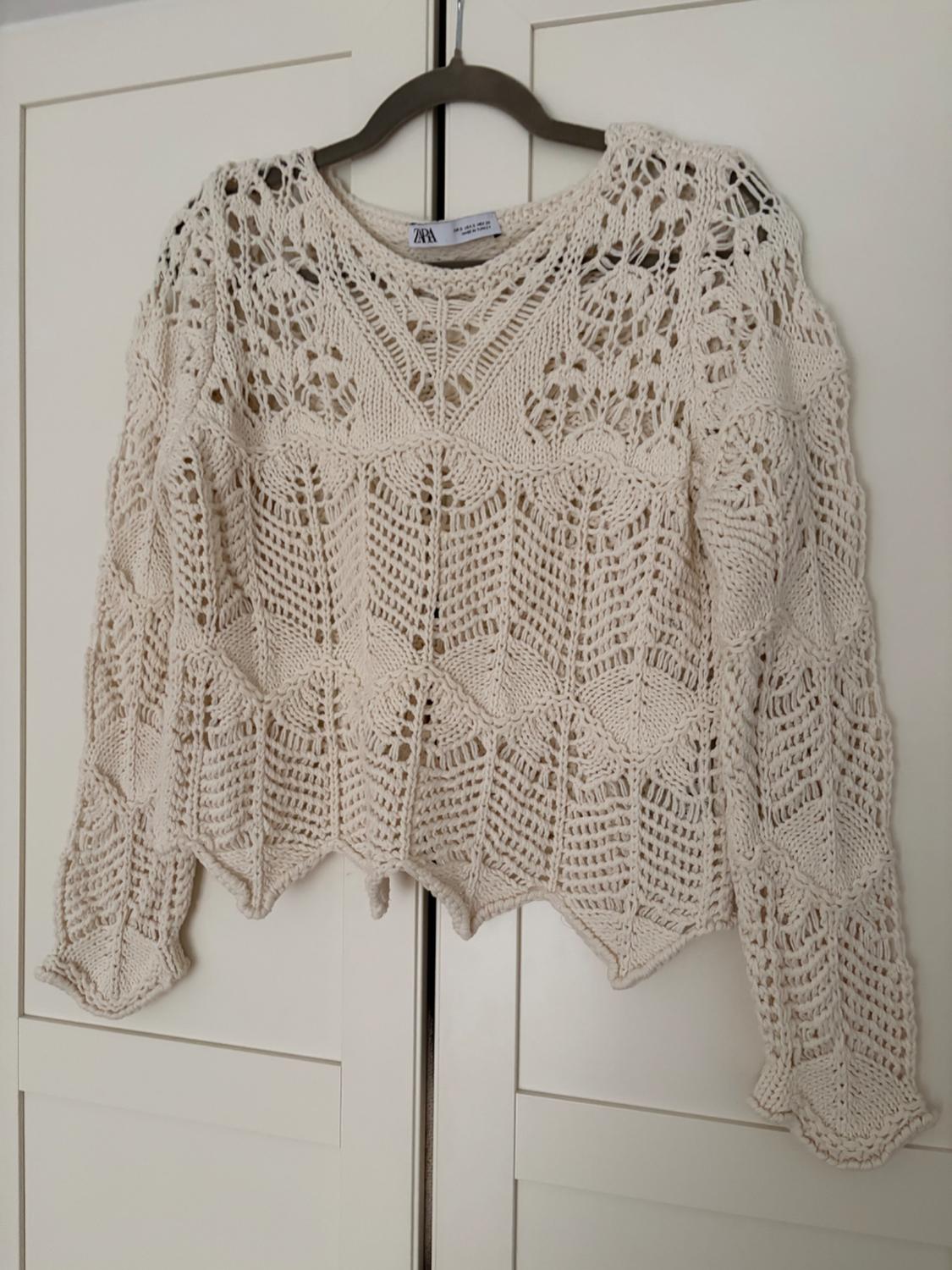 Beige crochet jumper hanging on a hanger in front of a white wardrobe. Openwork design with geometric and floral patterns.