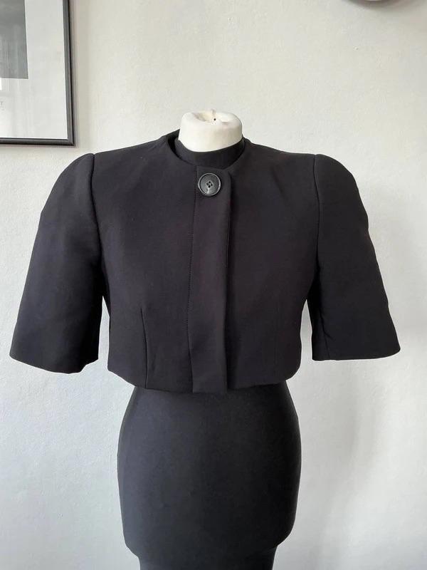 Mannequin wearing a short-sleeved black cropped jacket with a top button, over a black dress. White background with a grid.