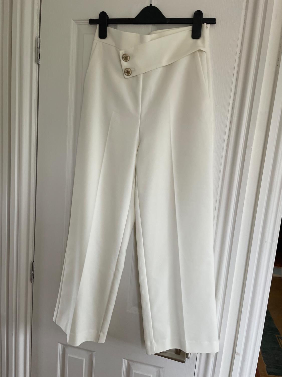 White, wide-leg trousers hanging on a black hanger, with two gold buttons on the waistband, over a white door.