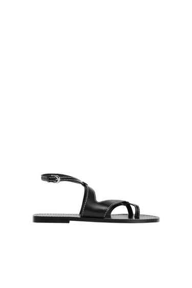 Black flat sandal with toe strap, ankle buckle fastening and white stitching, side view on a white background.