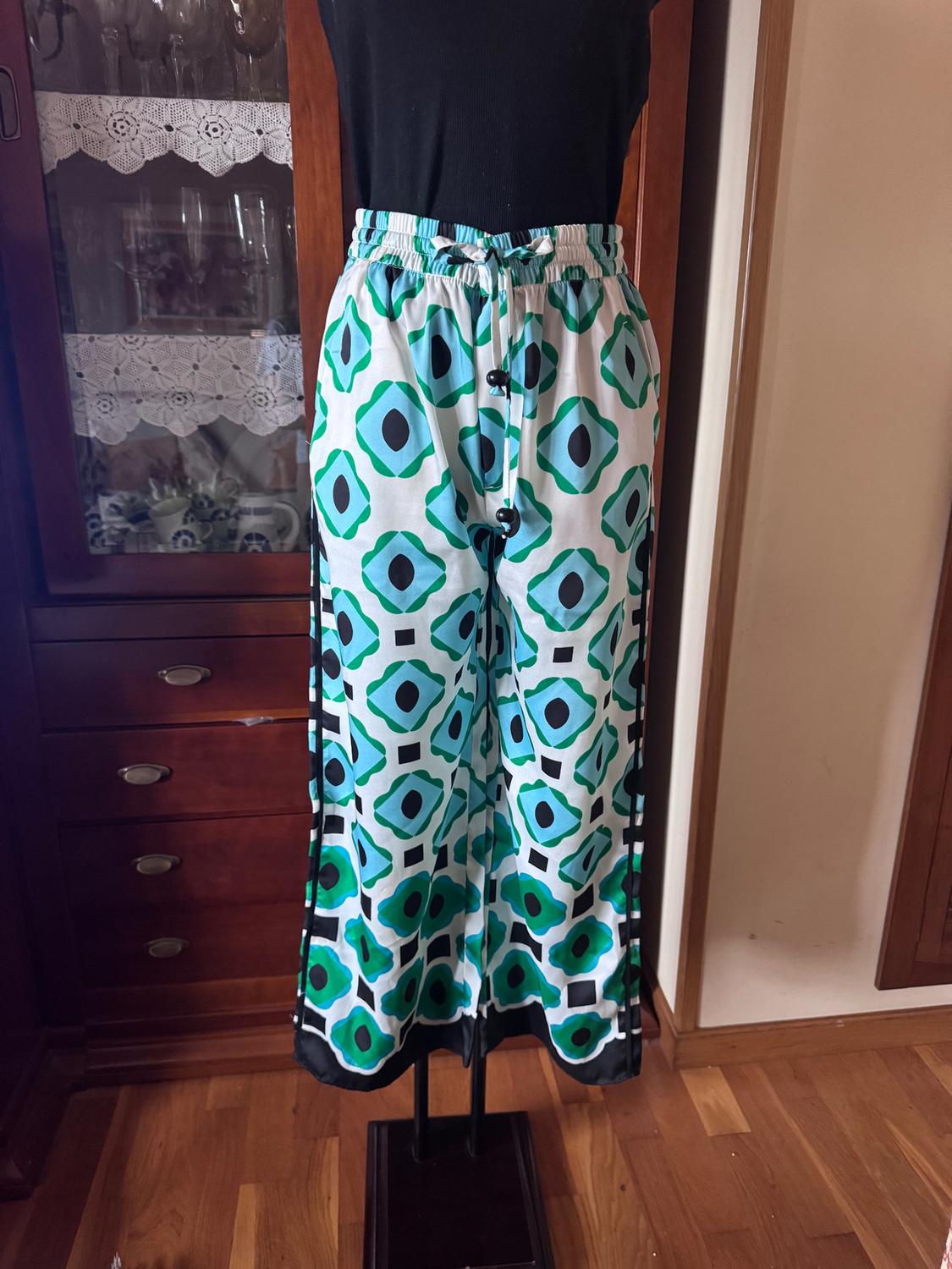 Mannequin with green and black geometric print trousers, white background, and black sleeveless top. Wooden furniture in the background.