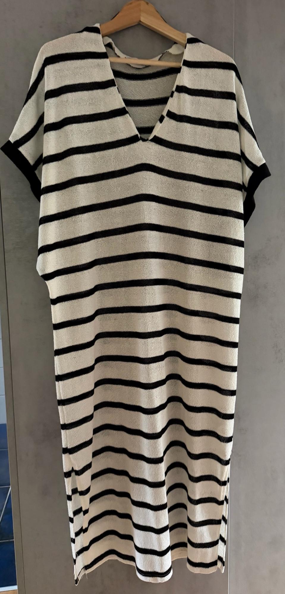 White knitted dress with black horizontal stripes and a V-neck, hanging on a wooden hanger against a grey wall.