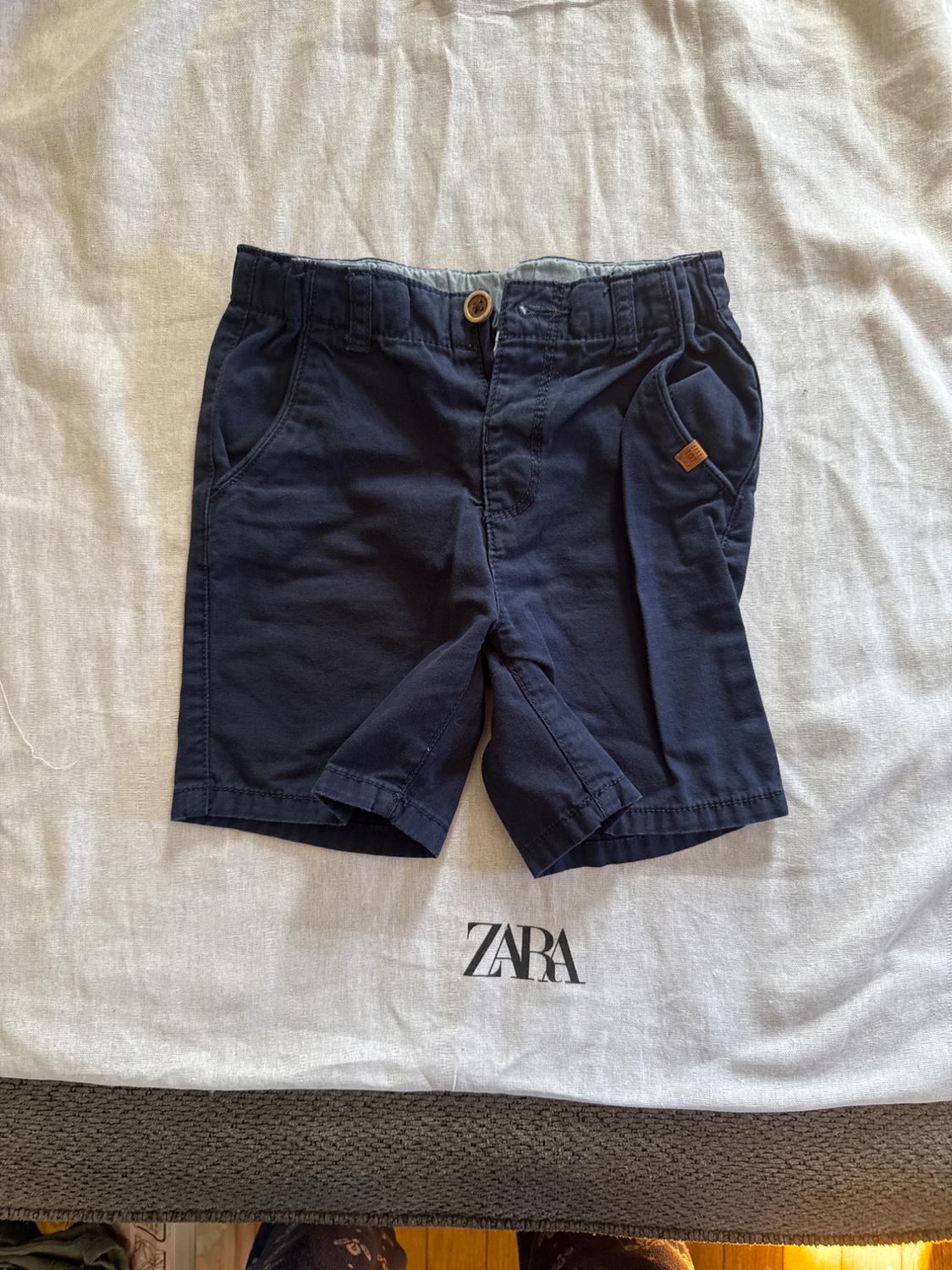 Navy shorts with a wooden button on white fabric. Visible text: "ZARA".