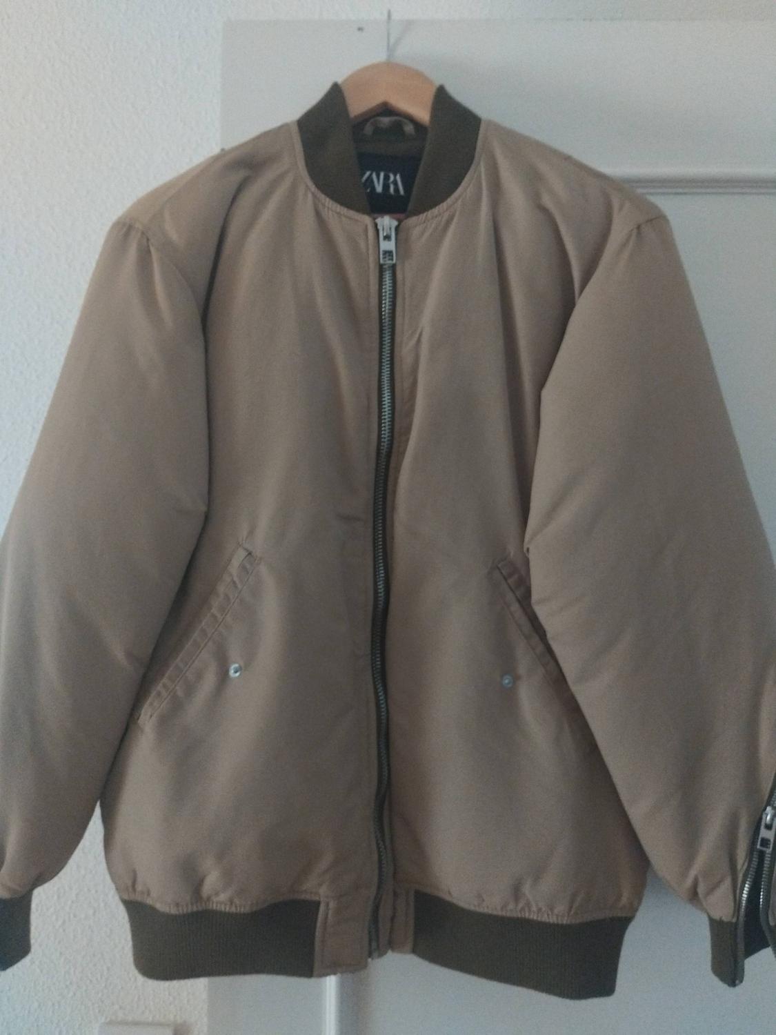 Beige bomber jacket hanging on a coat hanger. Design with a front zip, dark collar and cuffs. ZARA label visible inside.