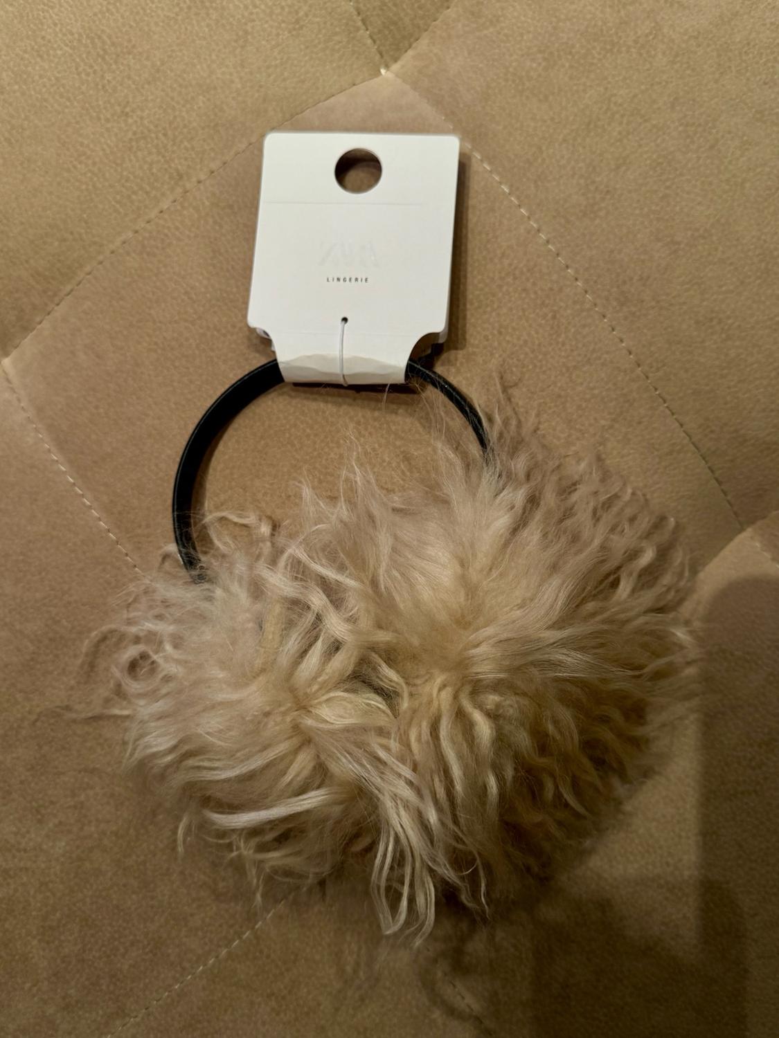 Black hair tie with a beige fur detail on a fabric background. Label bearing the text ZARA LINGERIE.