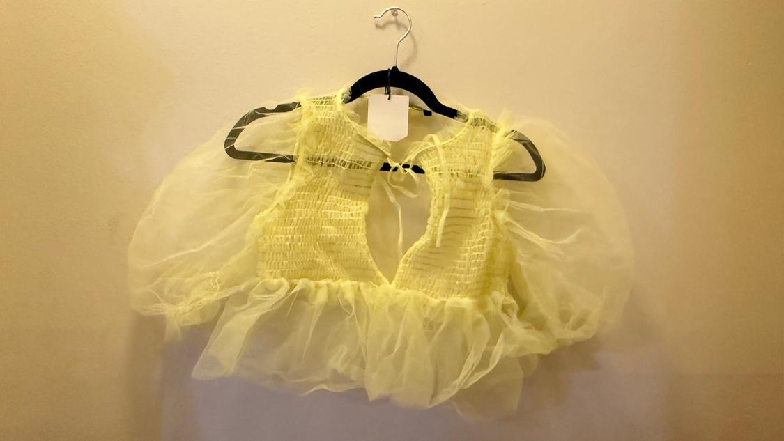 Yellow tulle blouse with puffed sleeves hanging on a black hanger against a beige wall.
