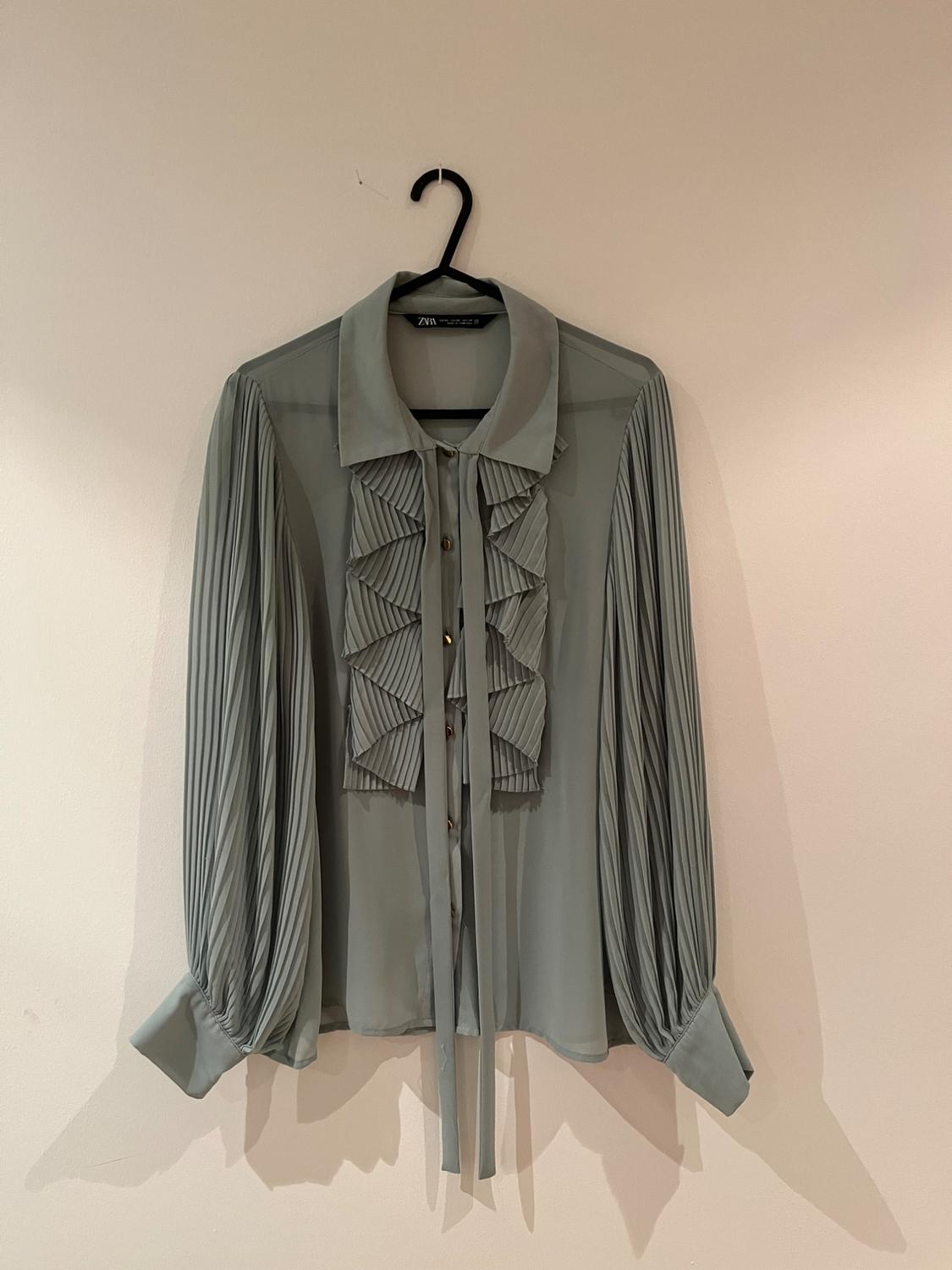 Light grey blouse with pleated long sleeves and ruffles on the front, hanging on a black hanger against a white wall.