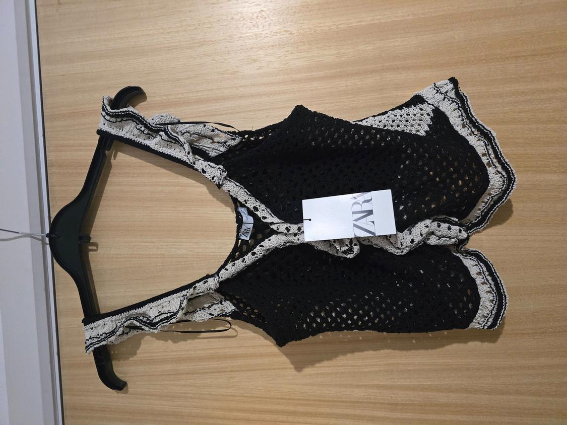 Black crochet top with white trim hanging on a black hanger against a light wood background. Visible label with "ZARA" text.