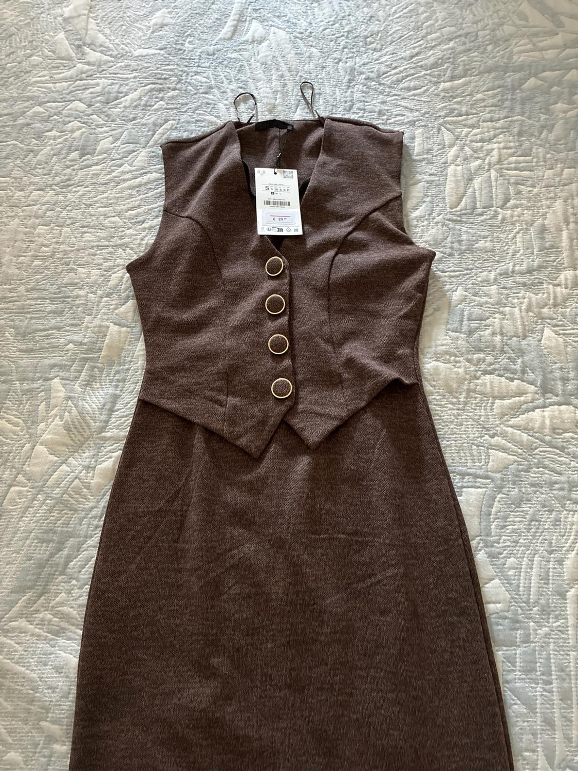 Brown sleeveless waistcoat-style dress with four buttons, laid out on a textured light-coloured bedspread.