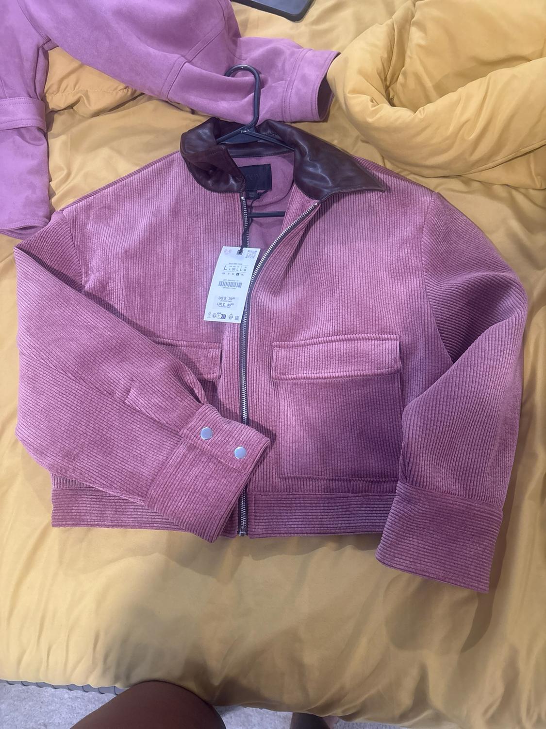 Pink corduroy jacket with a brown collar, zipper, and front pockets, with tags, on a bed with wrinkled yellow sheets.