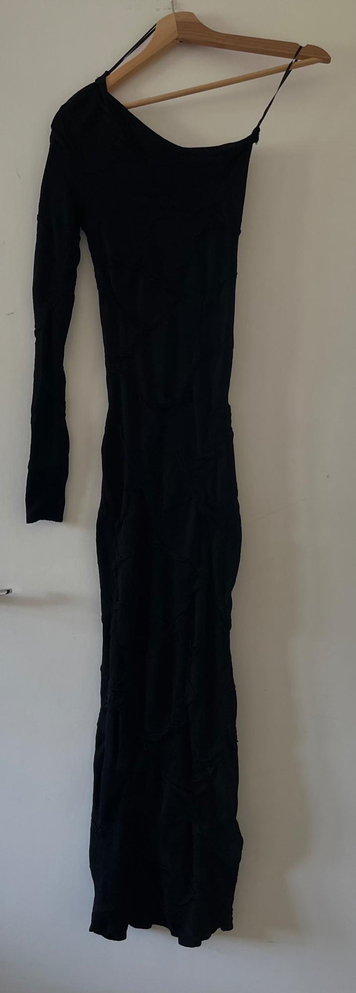 Textured asymmetric black maxi dress, with one long sleeve and a spaghetti strap, hanging on a wooden hanger against a white door.