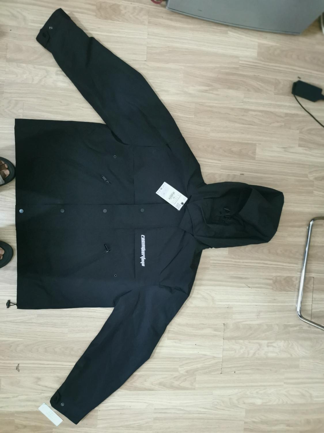 Black jacket with hood and press studs, laid out on a light wooden floor. Visible label on the chest. Text: "commuting wear".