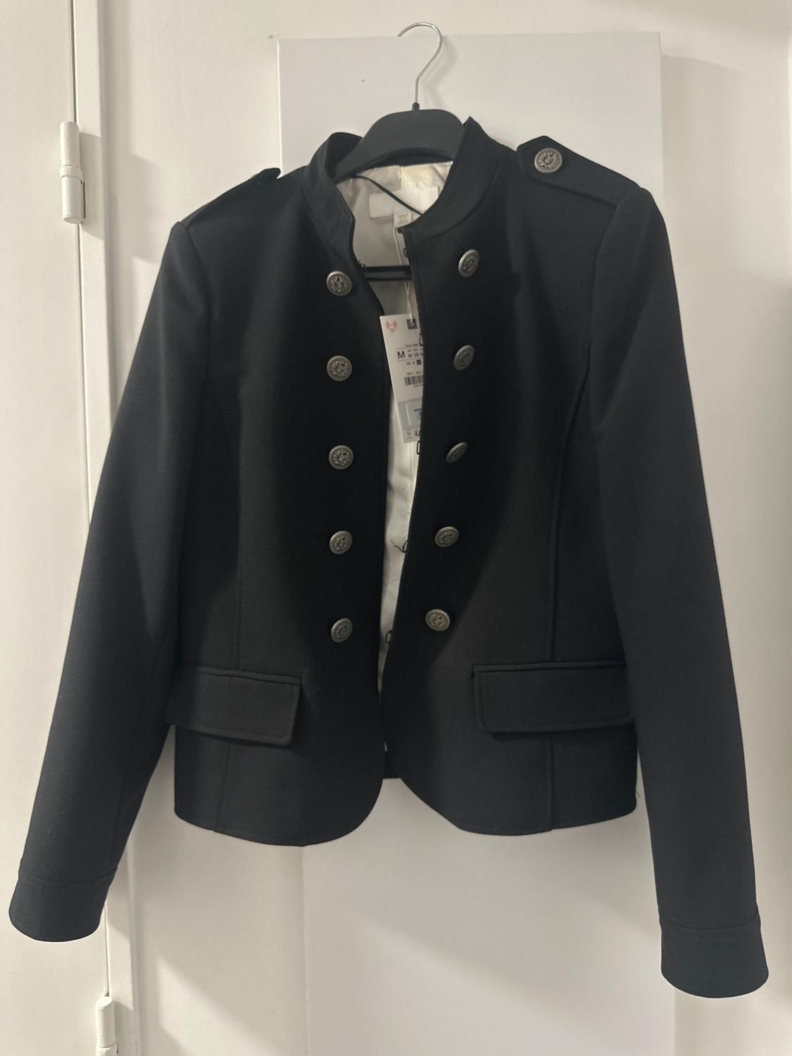 Black jacket with metal buttons and epaulettes, hanging from a hanger on a white door. Inside label with the text M.