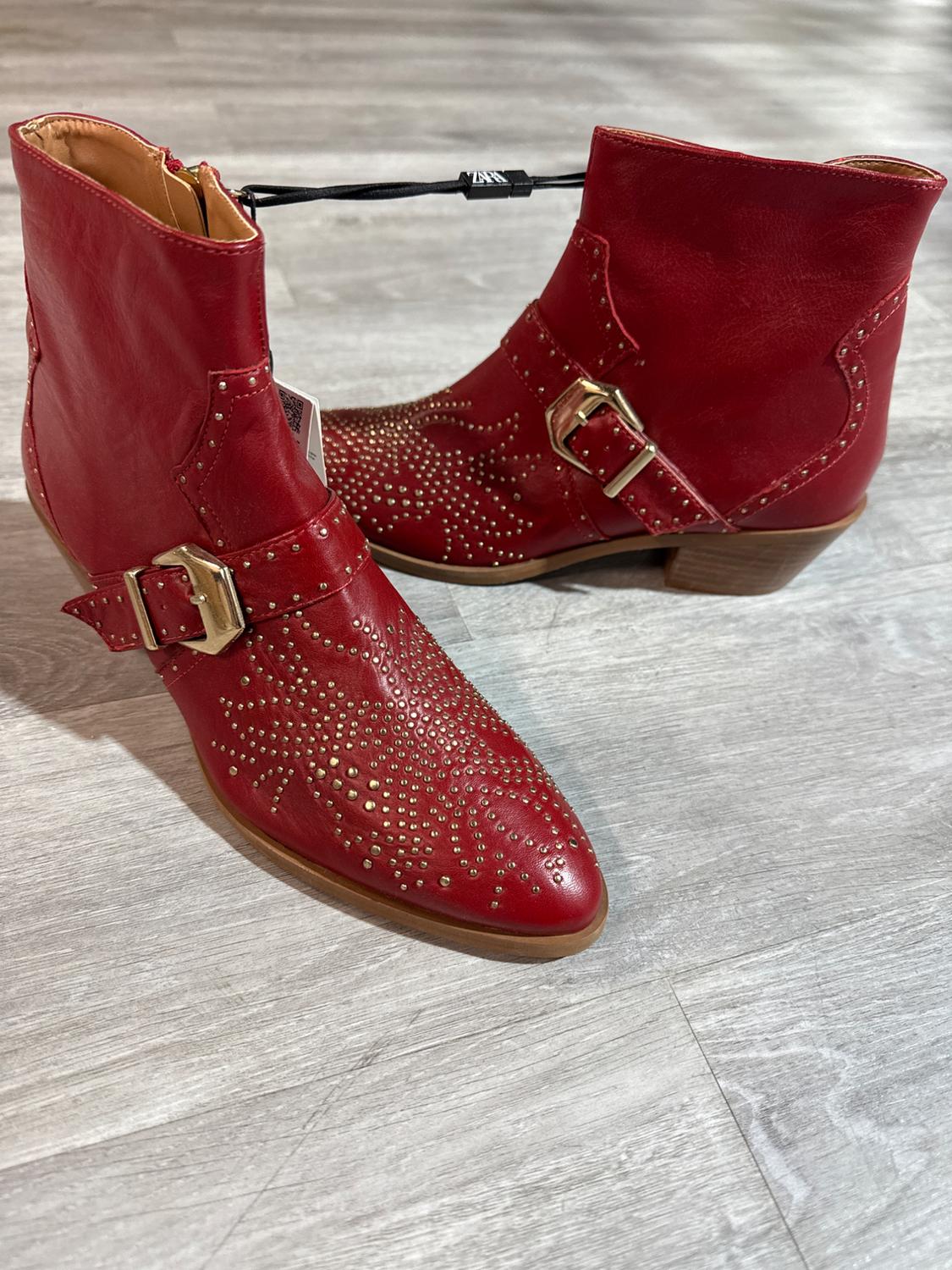 Red leather ankle boots with gold studs and buckles, on a grey wooden floor.