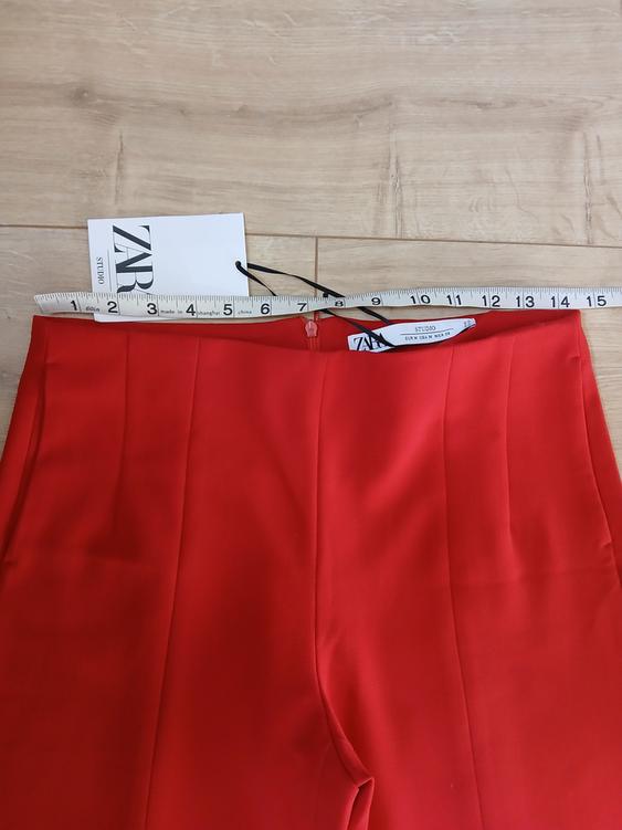 Red trousers on a wooden floor with a Zara Studio label. A tape measure is measuring the trousers' waist.