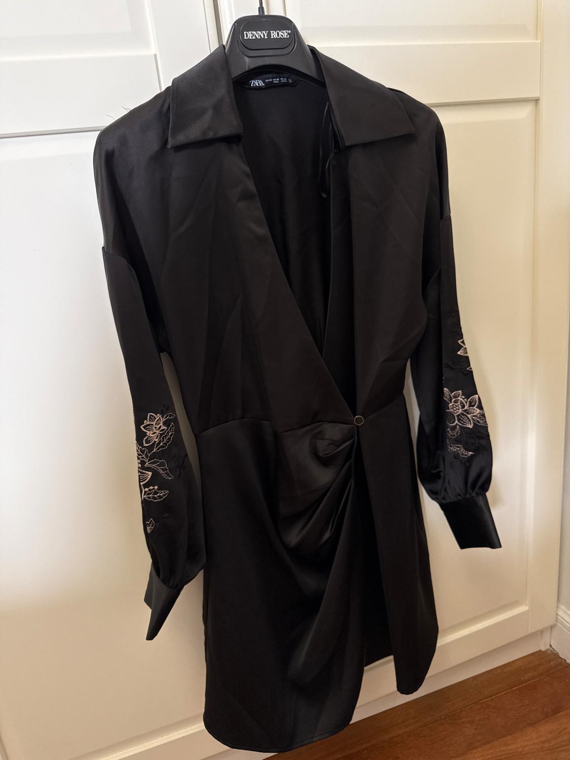 Black wrap dress with floral embroidery on sleeves, hanging on a hanger with DENNY ROSE text and ZARA tag, in front of a white wardrobe.