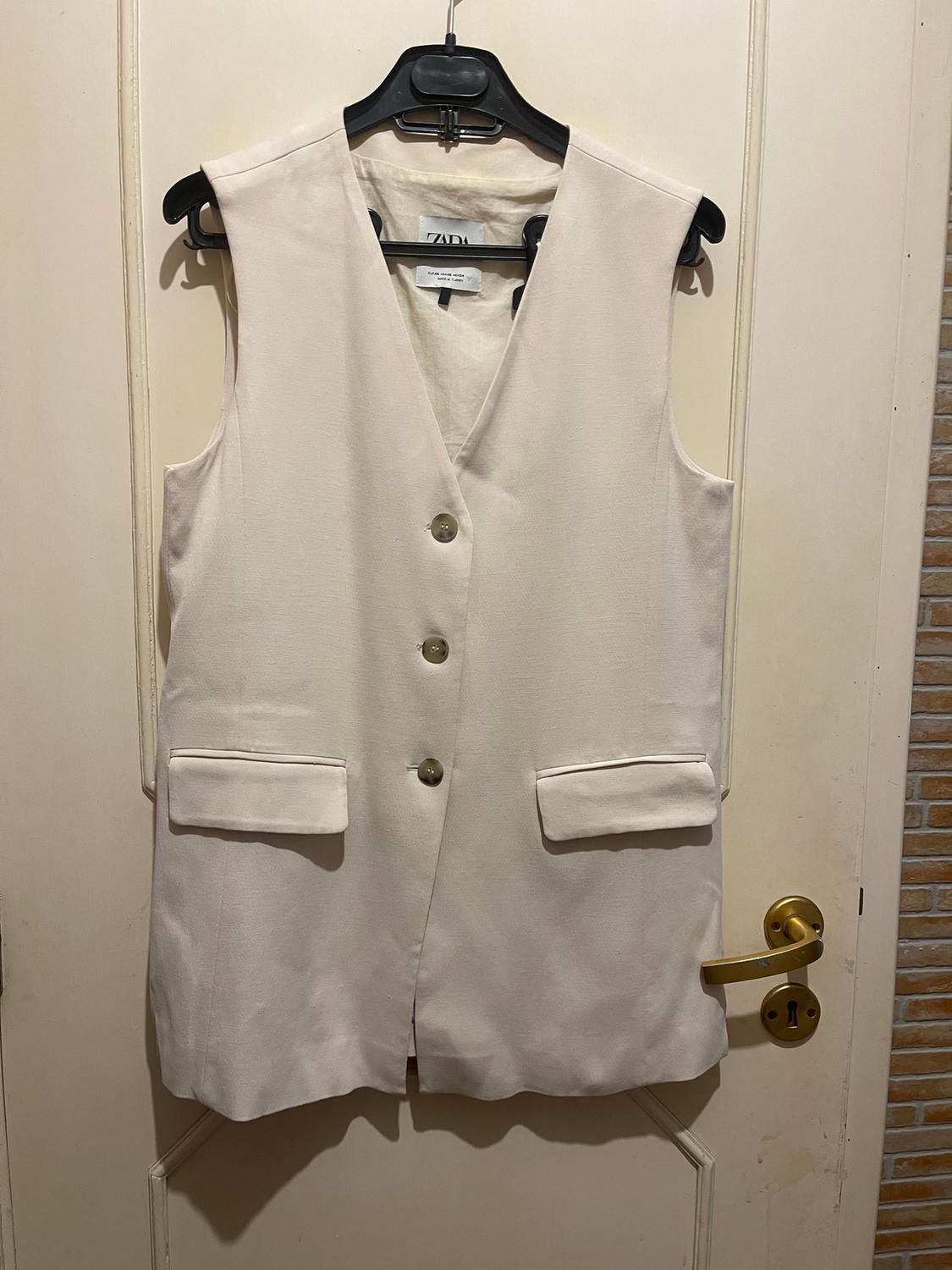Beige sleeveless waistcoat hanging on a black hanger over a door. It has three buttons and two front pockets. Visible tag.