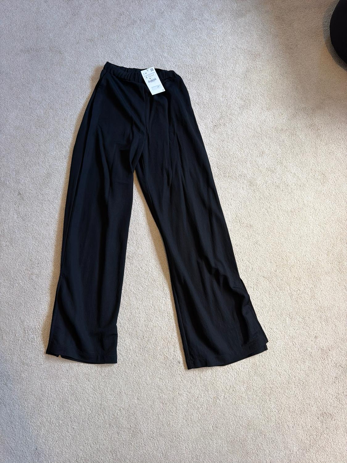 Flowing black wide-leg trousers with an elasticated waistband and white label, laid out on a light-coloured rug.
