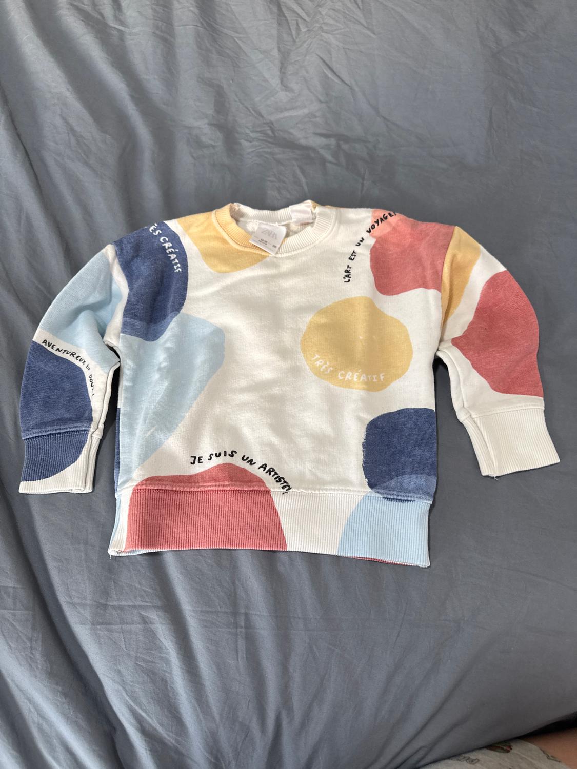 White sweatshirt with colourful paint splatters print and French text: "Je suis un artiste", "Très créatif", on a grey bed.
