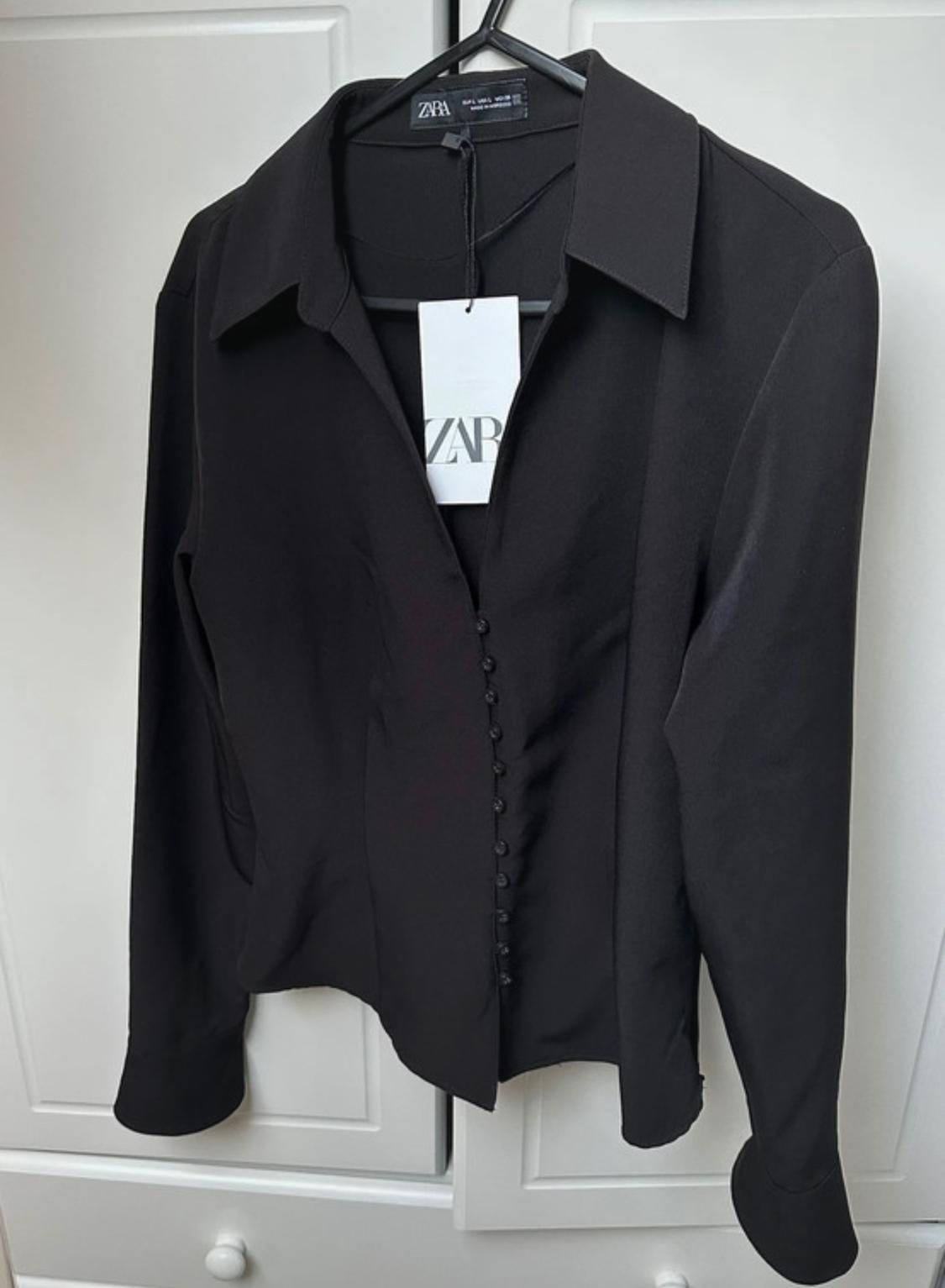 Black long-sleeved button-up shirt, hanging on a hanger. Visible label with the text "ZARA". White wardrobe background.