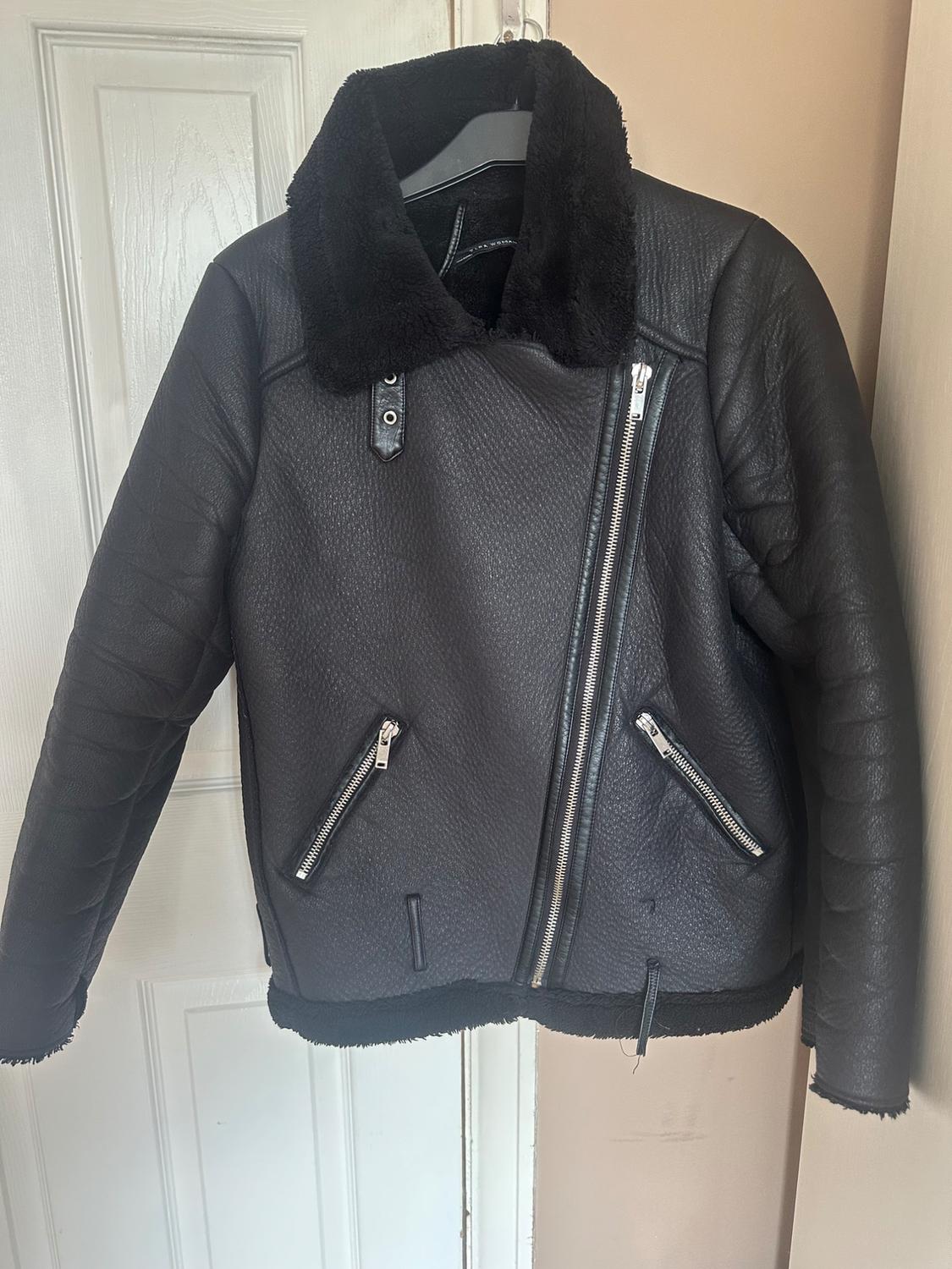 Black leather jacket with a diagonal zip and a faux fur collar, hanging on a hanger in front of a white door.