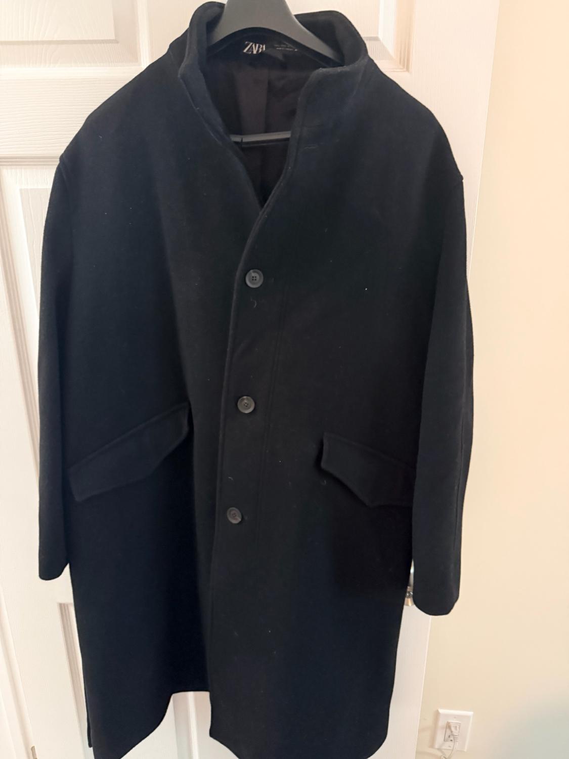 Black coat with buttons and pockets hanging on a hanger in front of a white door. Interior label with the text ZARA.