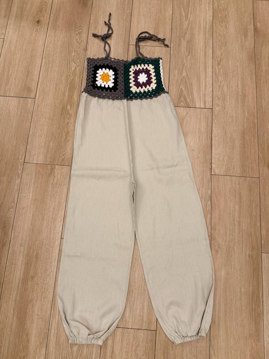 Jumpsuit with a crochet top and shoulder straps, and loose-fitting light beige trousers with ruched hems, on a wooden floor.