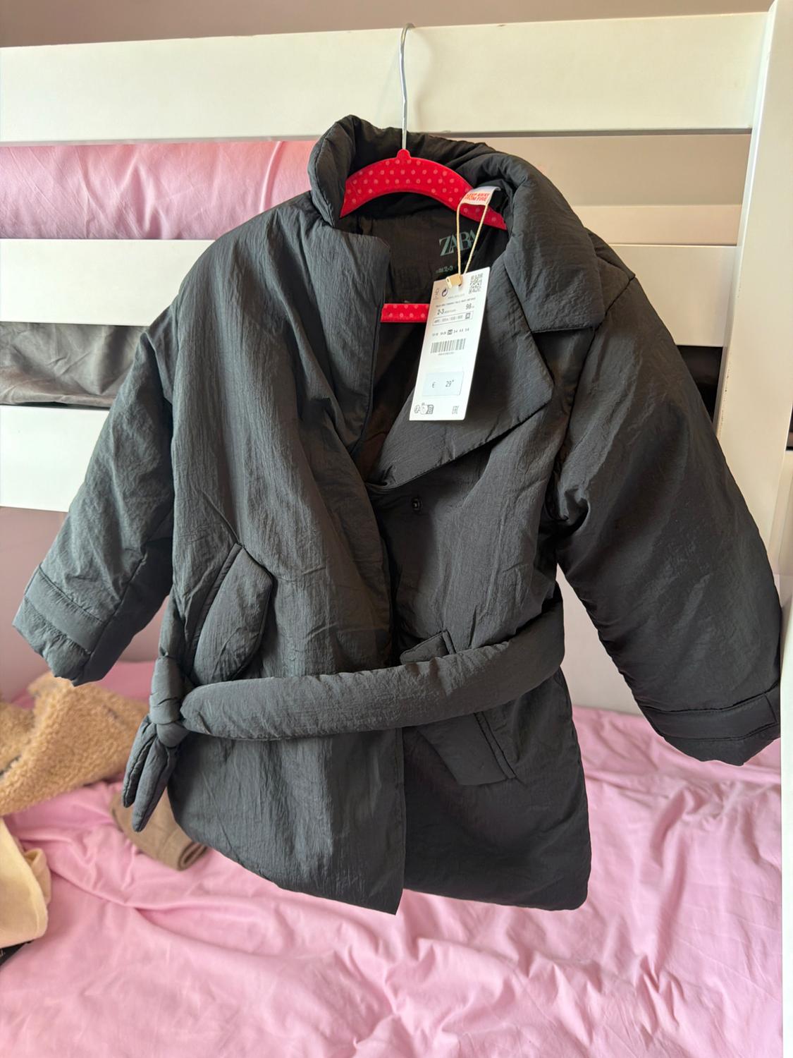 Black belted padded coat laid over a bed with pink sheets. Labels with the text ZARA and KEEP AWAY FROM FIRE.