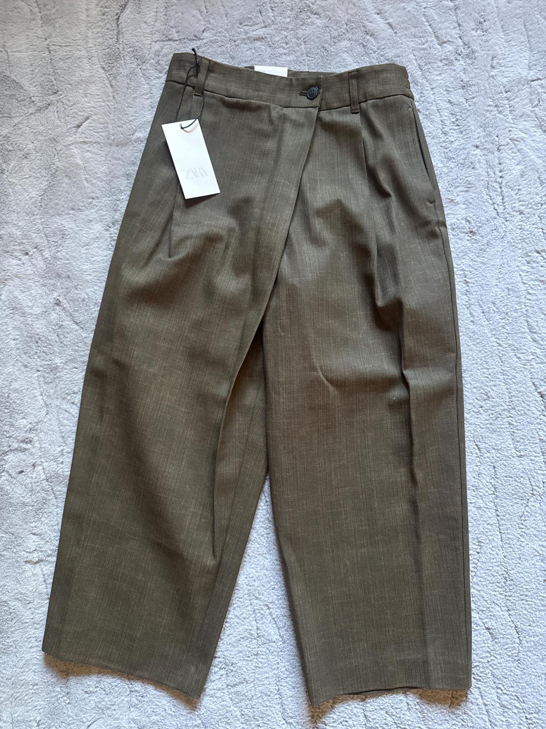 Brown pleated trousers with swing tag on a light grey textured background.
