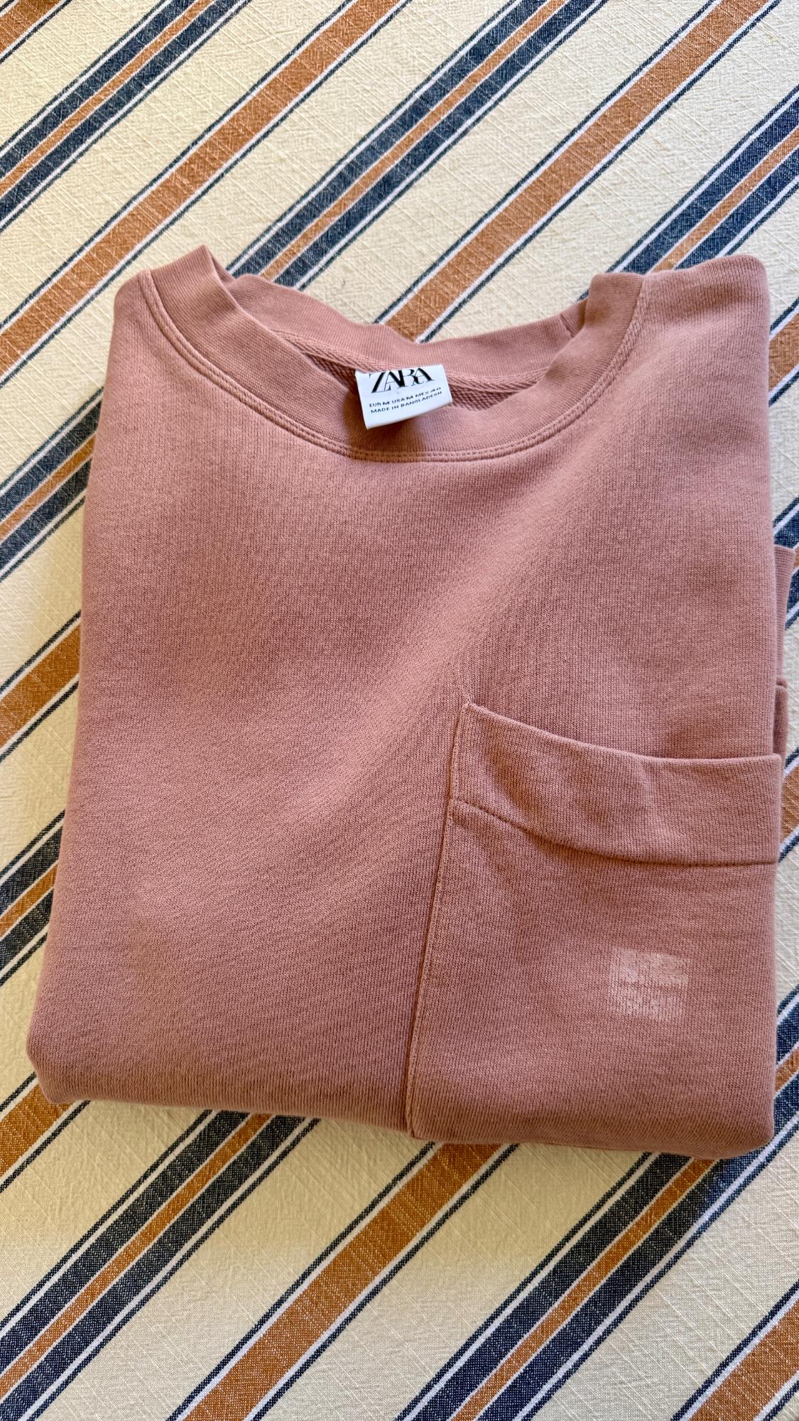 Folded pink sweatshirt with pocket on blue and mustard striped fabric. Text: ZARA EUR M USA M MEX 40 MADE IN BANGLADESH.