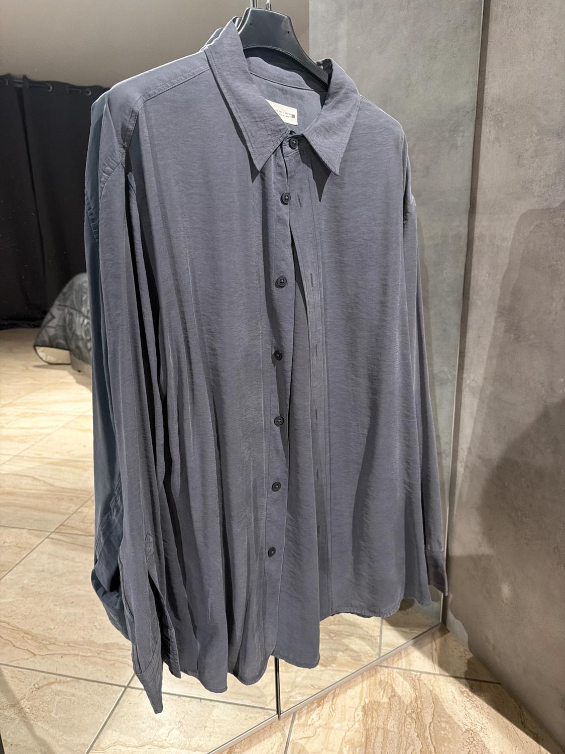 Grey long-sleeved shirt hanging on a black hanger, reflected in a mirror. Background with dark curtains and light tiled floor.