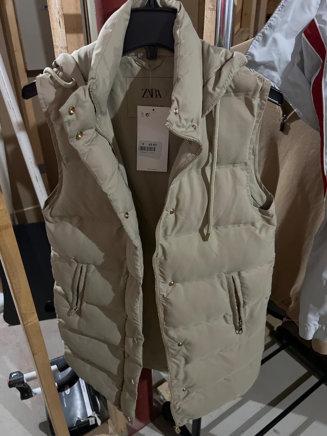 Front view of a beige puffer vest with a detachable hood and zippered side pockets.