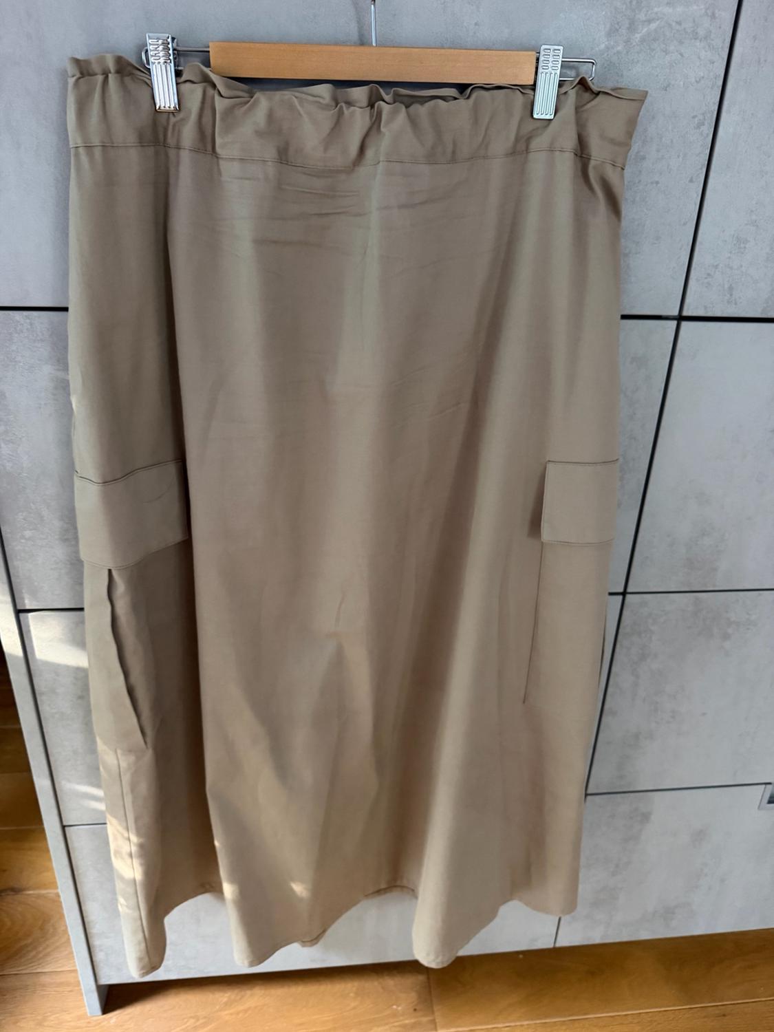 Beige cargo-style skirt with an elasticated waistband, hanging on a wooden hanger in front of a grey tiled wall.