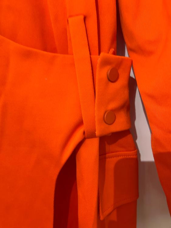 Detail of an orange garment with buttons and a belt of the same colour. The fabric has a ribbed texture and a visible pocket.