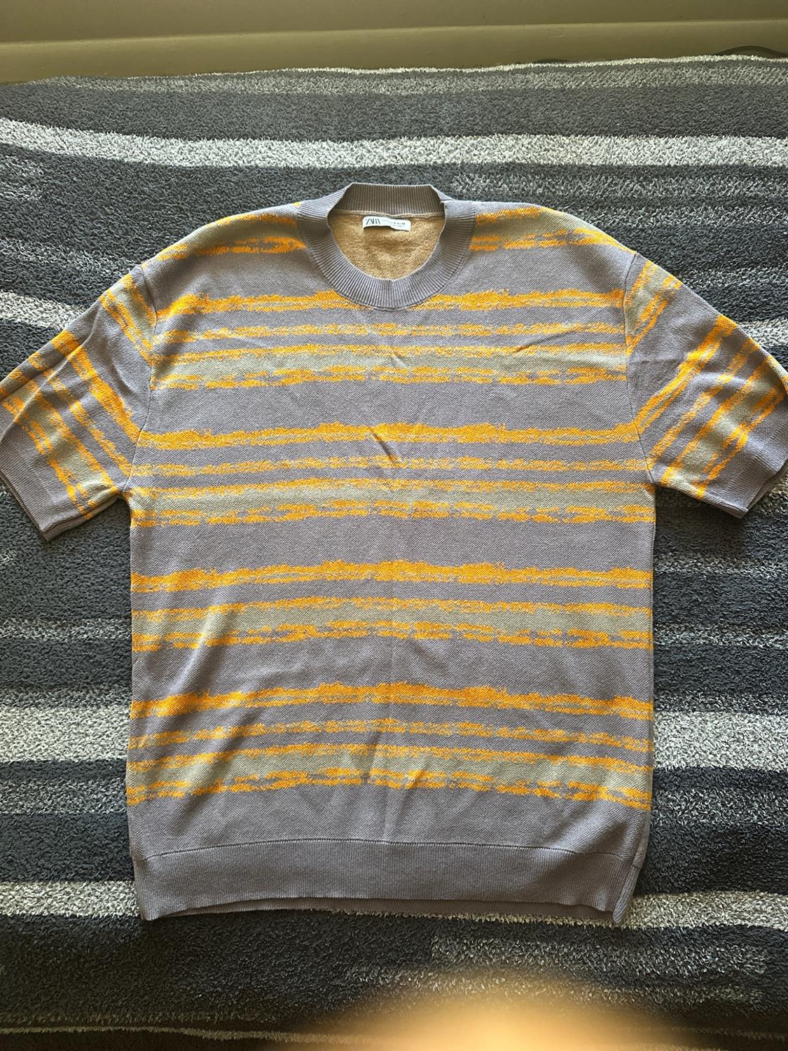 Gray knit T-shirt with short sleeves and yellow stripes over a surface with gray and white stripes. ZARA label at the neck.