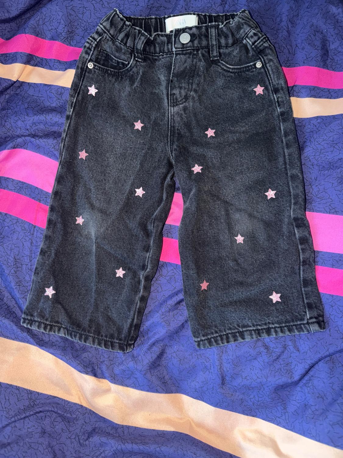 Black denim trousers with pink stars against a blue, pink, and orange striped background. Label with the text ZARA.